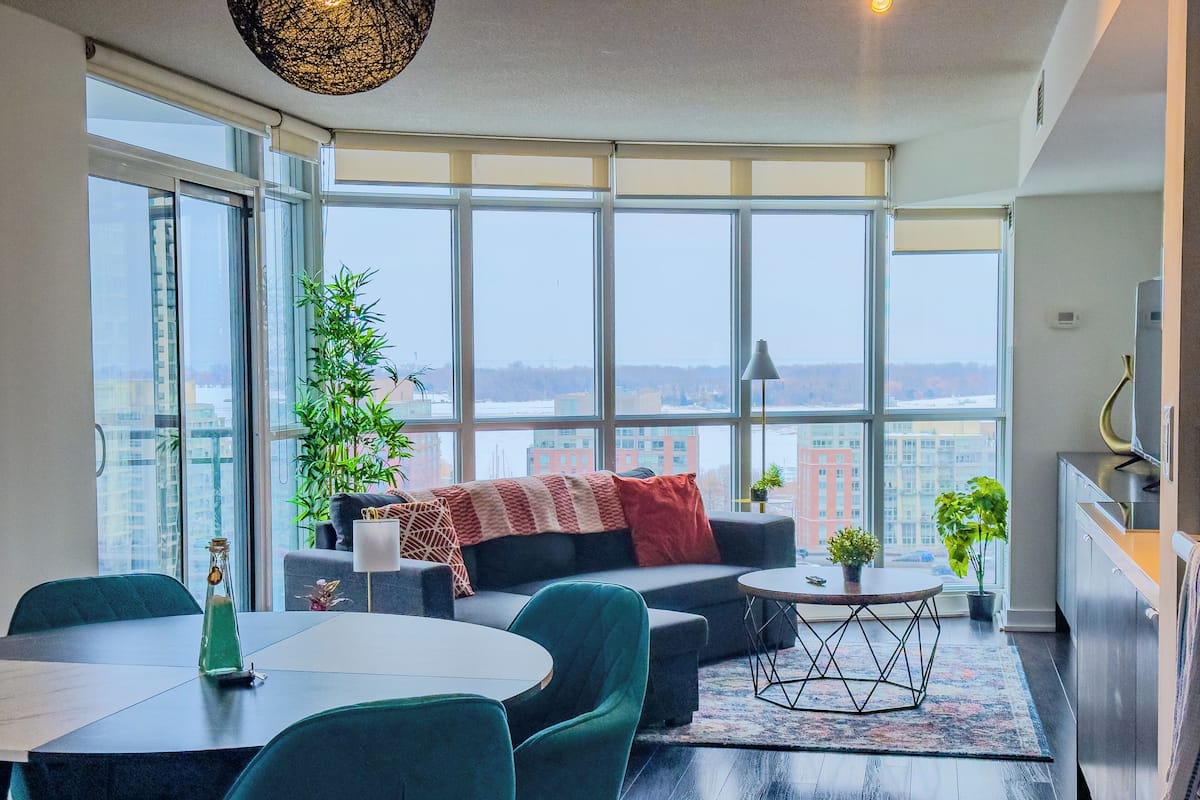A modern living room with a large window, a sofa, a dining table, and a kitchen area.