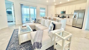 A modern living room with a sectional sofa, a glass coffee table, and a kitchen area with white cabinets and stainless steel appliances. - Centrally-located coach home. (Punta Gorda)