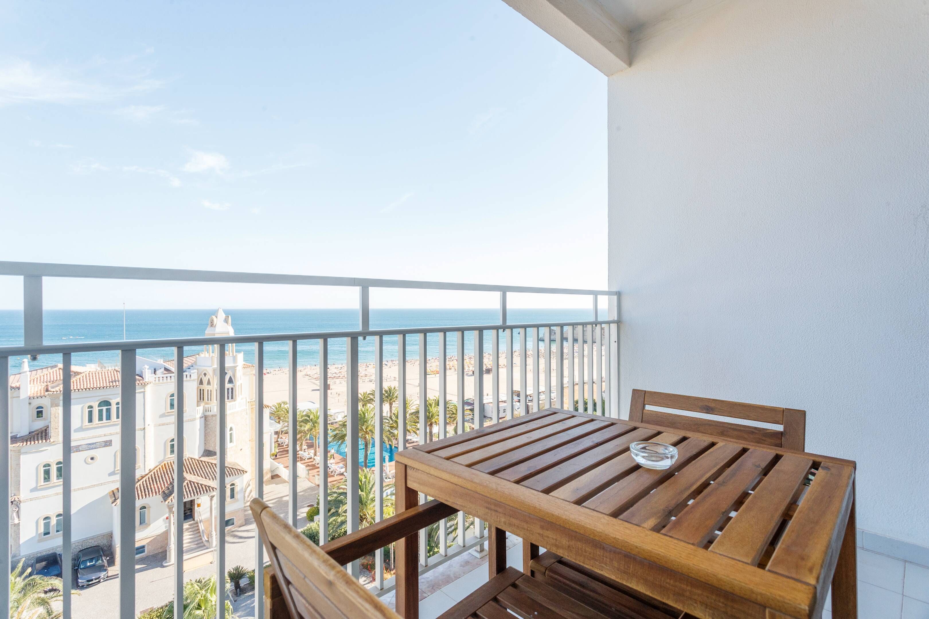 Ein Balkon mit Holztisch und Stühlen, mit Blick auf einen Strand und Gebäude.