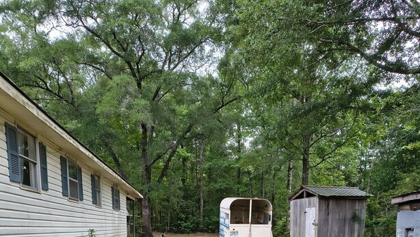 A white mobile home with a small attached shed, surrounded by trees and grassy area.
