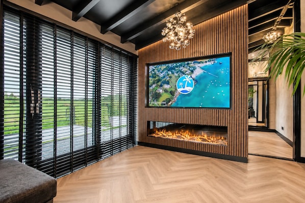 A modern living room with a large TV, wooden paneling, a fireplace, and large windows overlooking a green landscape.