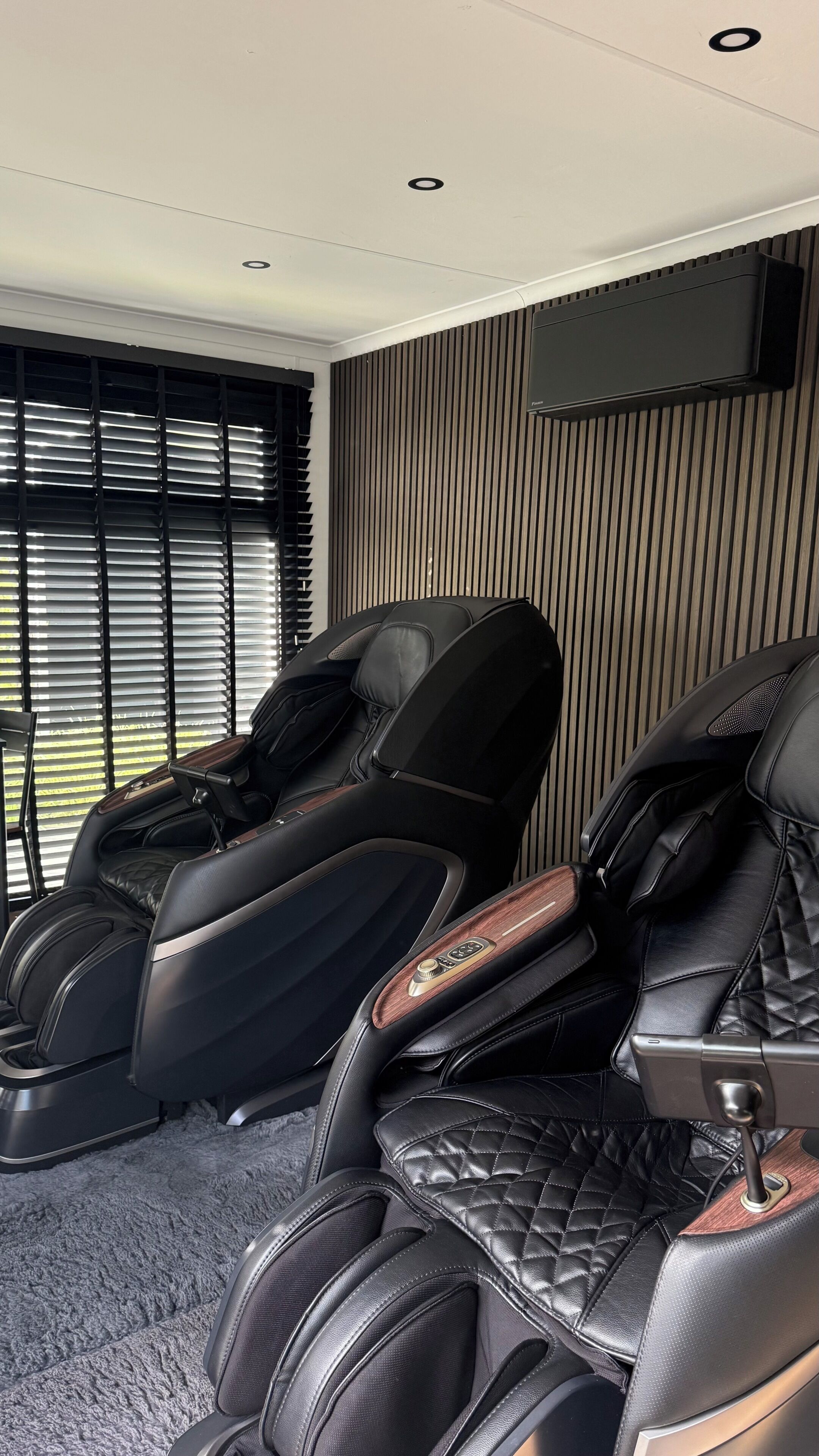 A massage room with two massage chairs, a flat-screen TV mounted on the wall, and a window with blinds.