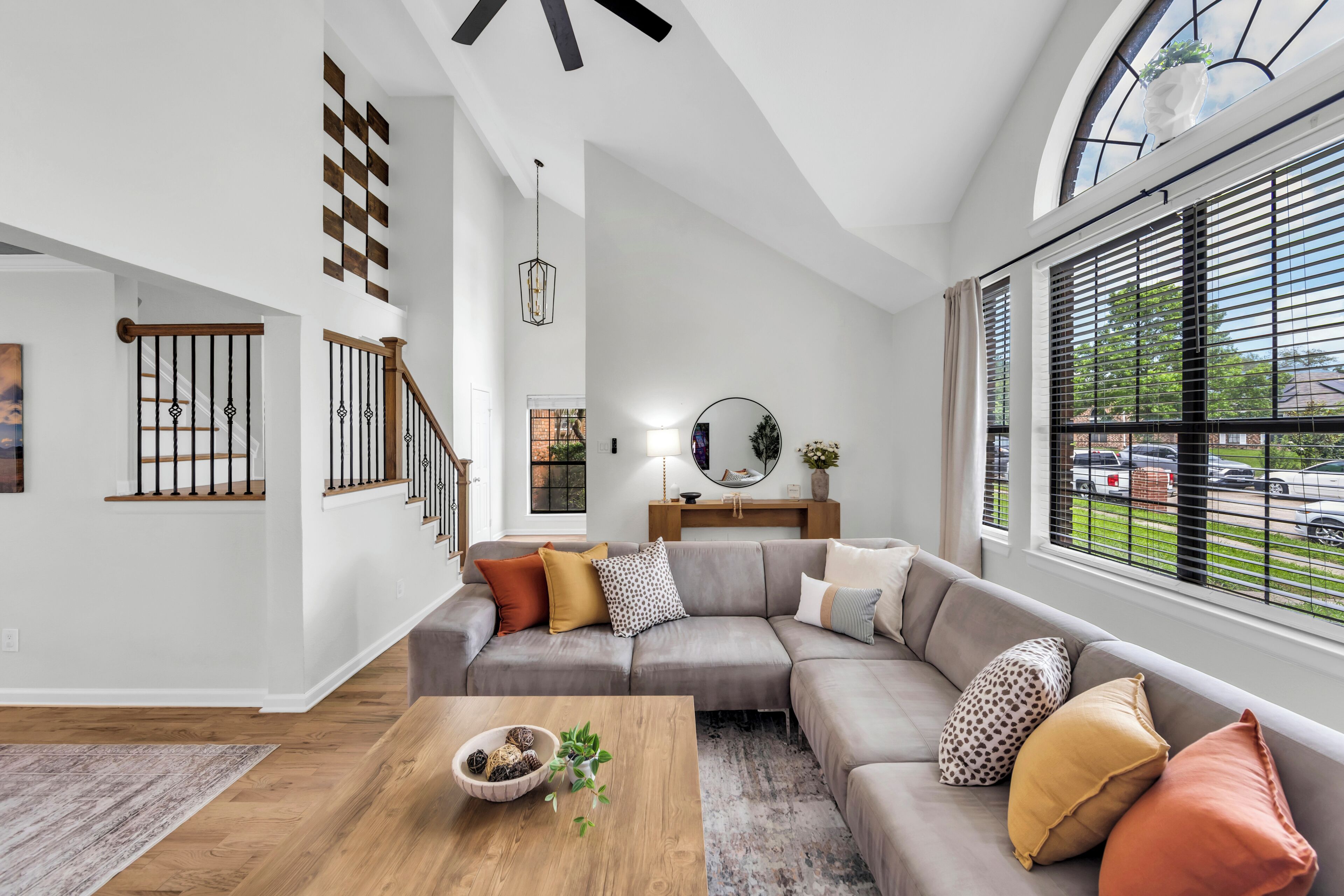 A modern living room with a wooden staircase, a sectional sofa, a coffee table with decorative items, and large windows with blinds.
