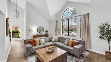 A modern living room with a sectional sofa, a wooden coffee table, a large window with shutters, and a potted plant.