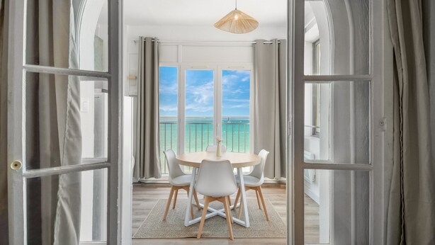 Eine offene Tür führt in einen Essbereich mit Tisch und Stühlen, einer Hängelampe und Meerblick durch große Fenster.