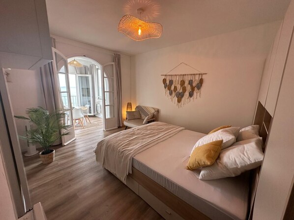 Une chambre Ă coucher avec un lit, une table de chevet, une lampe, des Ă©lĂ©ments de dĂ©coration murale et une vue sur lâextĂ©rieur, visible Ă travers une porte ouverte.