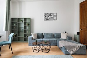 En stue med en grå sofa, to runde borde, et grønt reolsystem og en trædør.