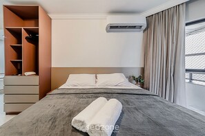 Ein modernes Hotelzimmer mit Bett, Einbauschrank, Klimaanlage und Blick durch ein mit Vorhängen versehenes Fenster.