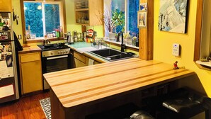 A kitchen with a wooden countertop, stove, sink, and hanging pots. There are windows with views of trees, and a painting on the wall. - Beautiful, private  2-bedroom cabin only a few miles from downtown Talkeetna (Talkeetna)