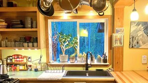 A kitchen with wooden cabinets, a stove, hanging pots, a window with a view of trees, and a patterned rug. - Beautiful, private  2-bedroom cabin only a few miles from downtown Talkeetna (Talkeetna)