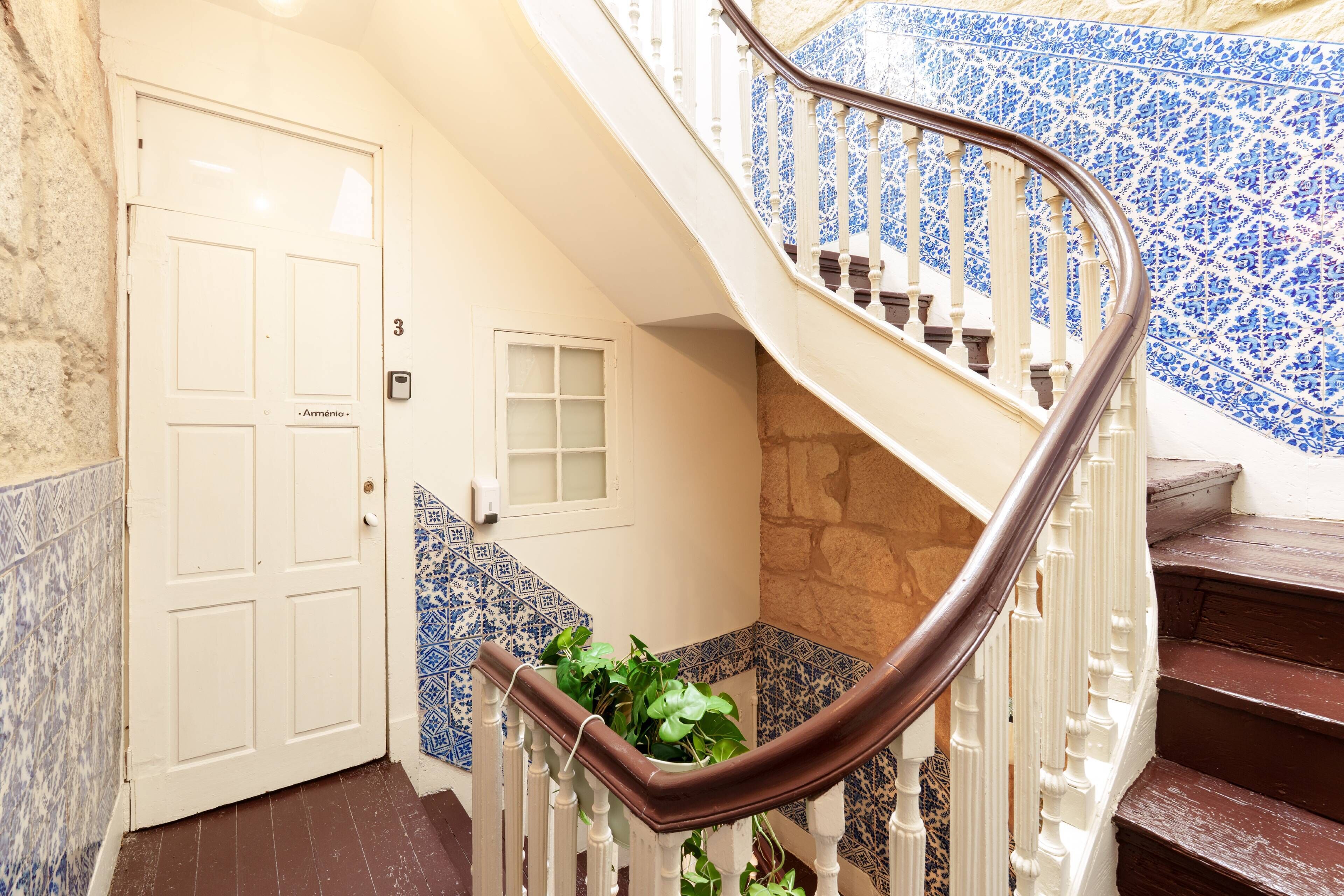 A spiral staircase with white balusters and a brown handrail, leading to a room with a tiled wall and a door labeled 'Arménio'.