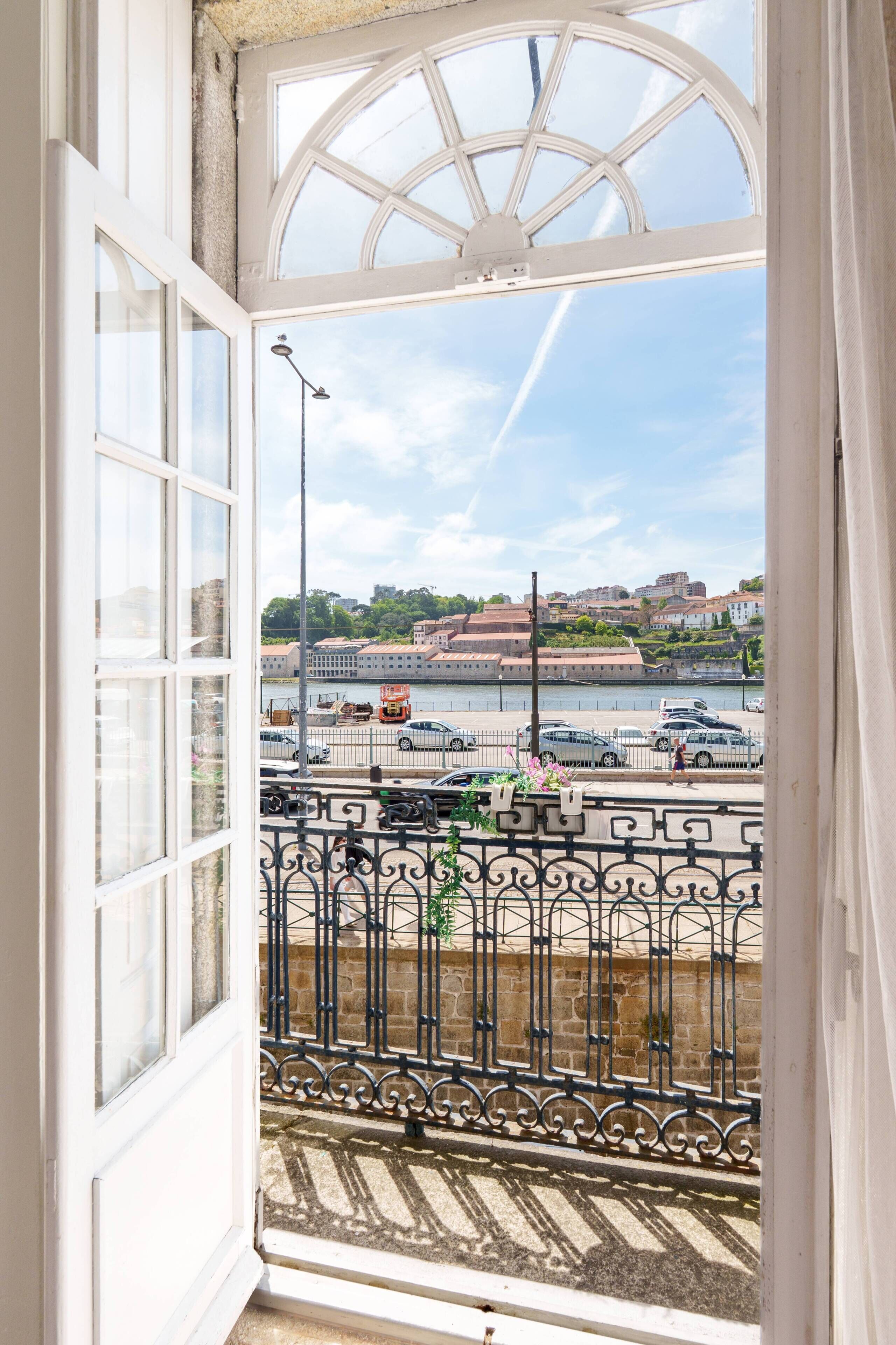 A view from a window with a white frame and a glass door, overlooking a street with a river, buildings, and a bridge.