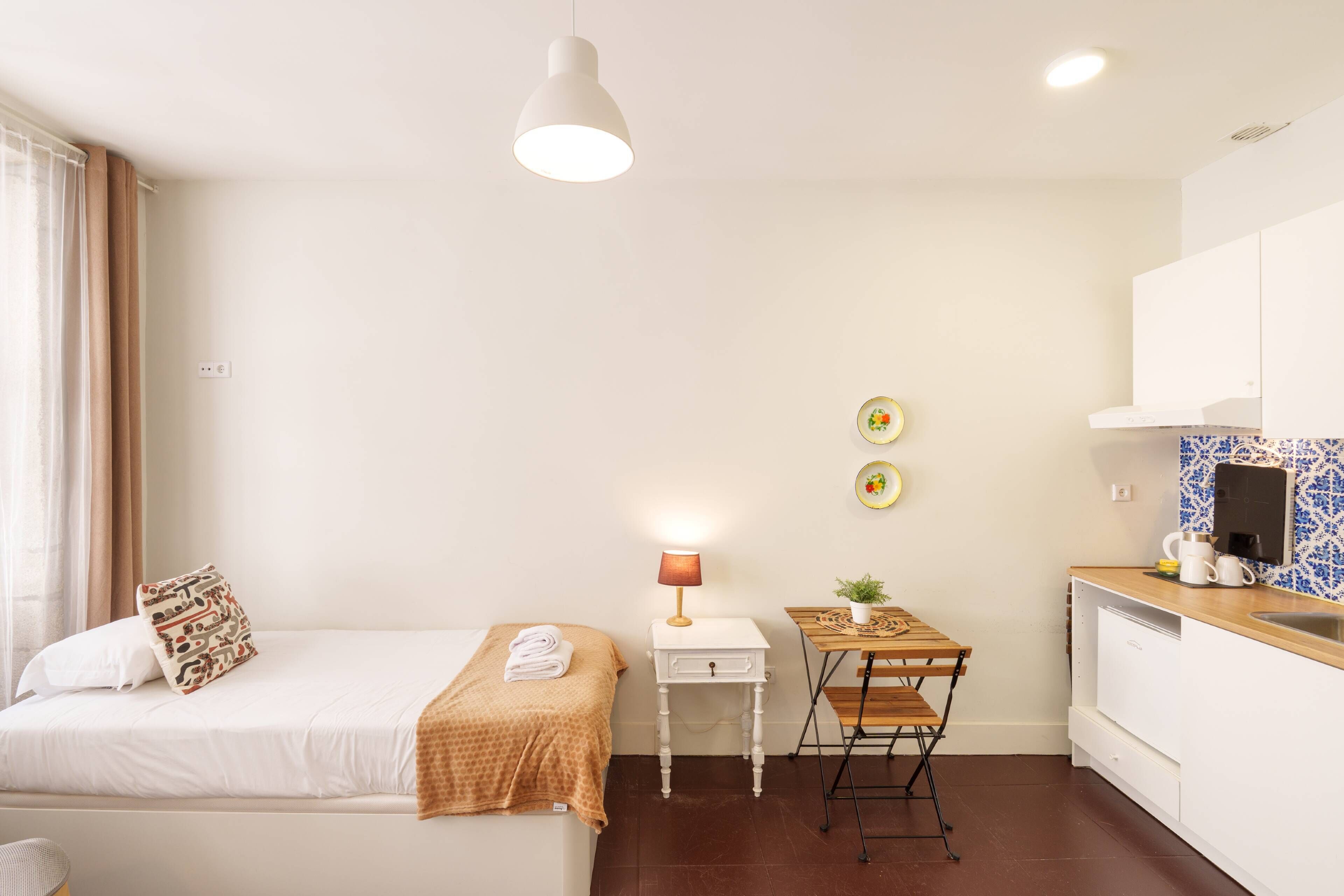 Un dormitorio con cama, mesita de noche, lámpara y una pequeña cocina con fregadero y encimera.