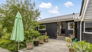 Un terrazza in legno con vasi di piante, un ombrello verde e una casa con facciata scura e tetto in tegole.