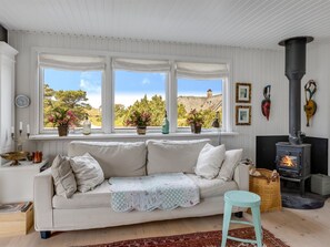 Ein gemütliches Wohnzimmer mit einem weißen Sofa, einem kleinen Tisch, einem Kamin und Blick ins Freie durch große Fenster.