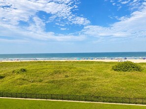 Une zone herbeuse avec un sentier et une clôture, qui mène à une plage où se trouvent des gens et des parasols.