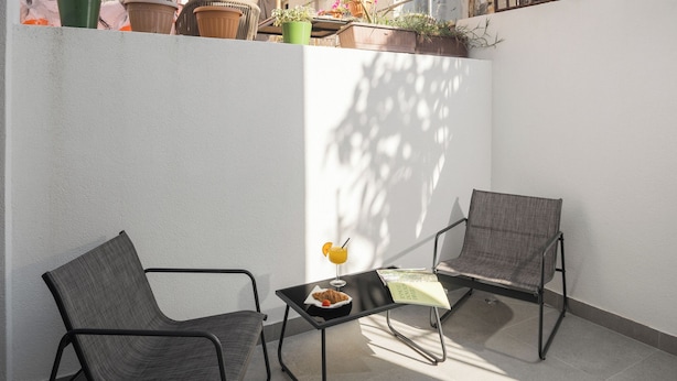 Un pequeño espacio al aire libre con dos sillas, una mesa, una planta en maceta y un balcón con plantas en macetas.