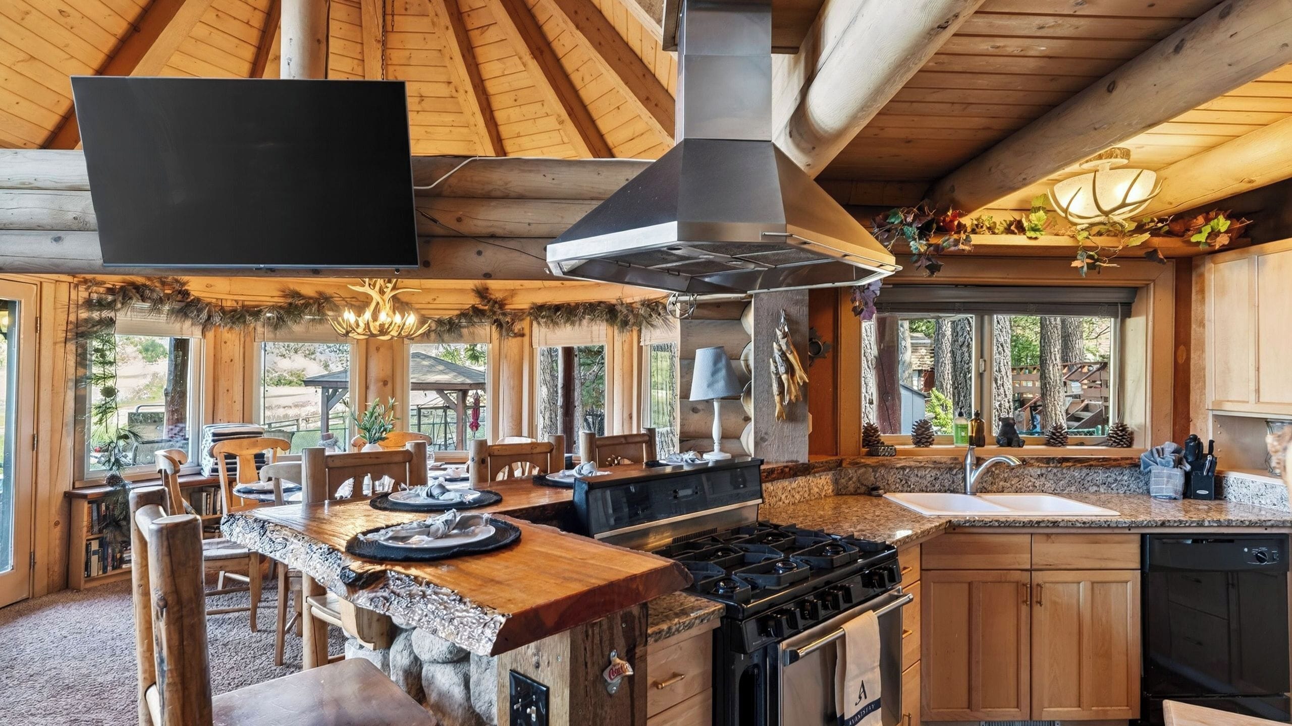 A rustic kitchen with a stone countertop, wooden cabinets, a gas stove, and a large window overlooking a forested area.
