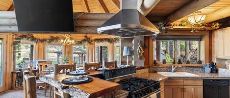 A rustic kitchen with a stone countertop, wooden cabinets, a gas stove, and a large window overlooking a forested area.