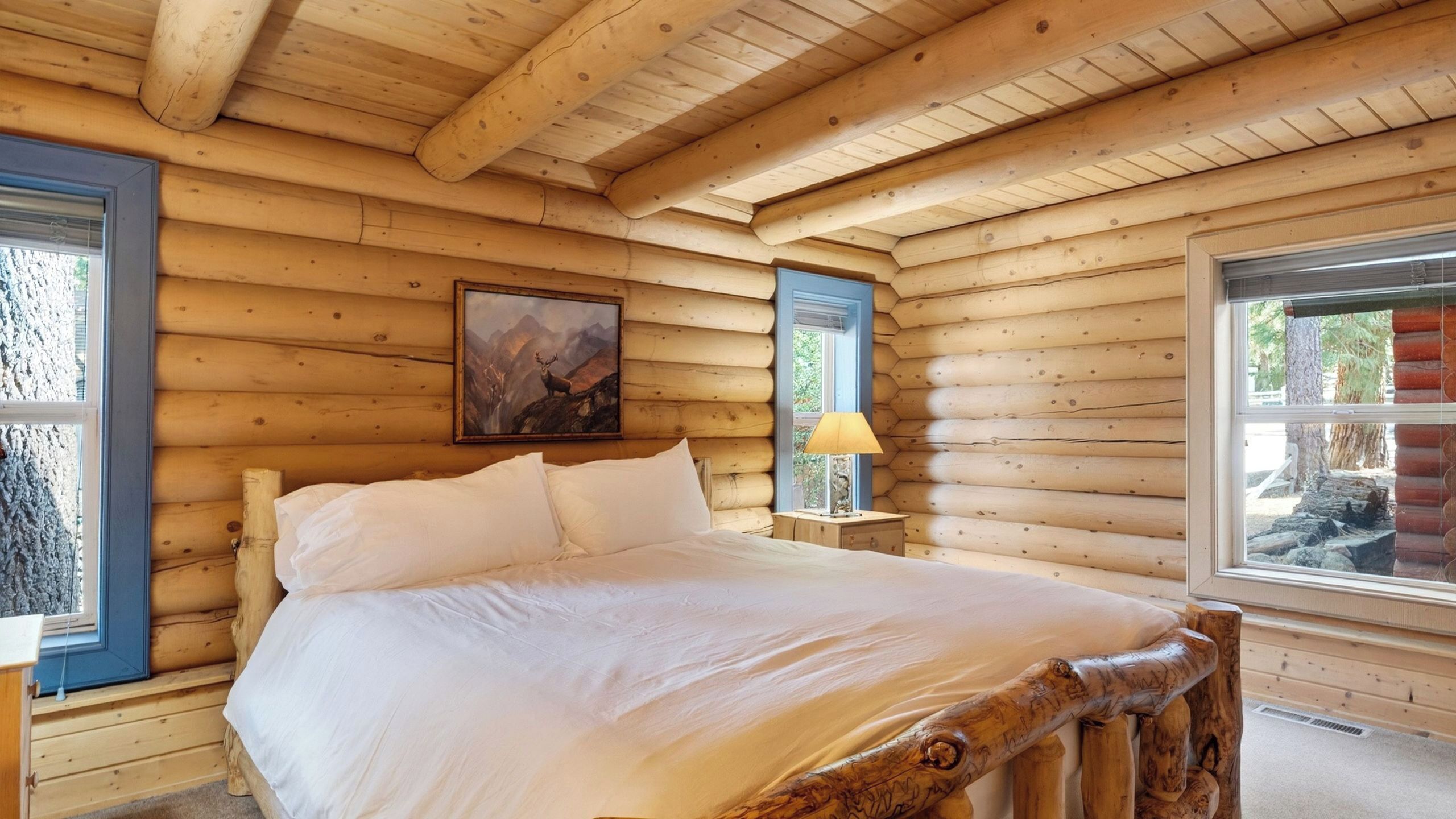 A wooden cabin bedroom with a bed, a painting, a nightstand, and a window.