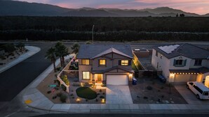 A suburban two-story house with a garage, a driveway, and a well-maintained front yard with palm trees and a small lawn. - La Casita Retreat Coachella by Avantstay Pool + Spa Desert Home + Putting Green + Game Room (Coachella)