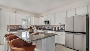 A modern kitchen with white cabinets, a granite countertop, a central island with seating, a built-in microwave, an oven, and a refrigerator. - La Casita Retreat Coachella by Avantstay Pool + Spa Desert Home + Putting Green + Game Room (Coachella)