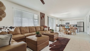 A modern living room with a sectional sofa, a dining area with a wooden table and chairs, and a kitchen with white cabinets and appliances. - La Casita Retreat Coachella by Avantstay Pool + Spa Desert Home + Putting Green + Game Room (Coachella)