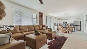 Uma sala de estar moderna com um sofá seccional, uma área de jantar com mesa e cadeiras de madeira, e uma cozinha com armários e eletrodomésticos brancos.