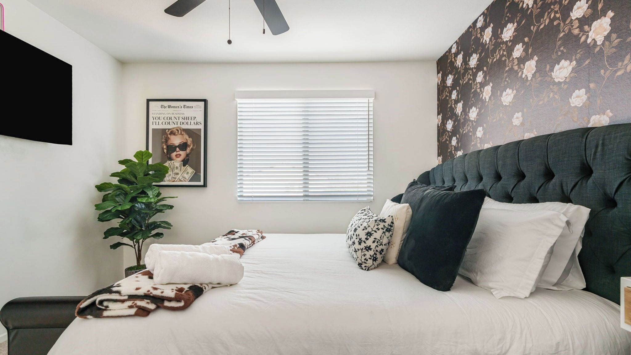 A bedroom with a large bed, a ceiling fan, a window with blinds, a wall-mounted TV, and a poster.