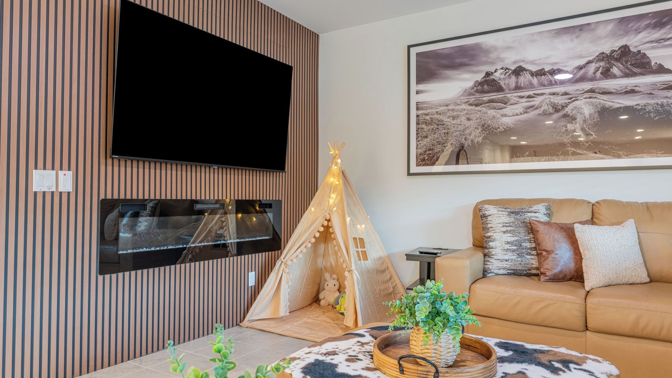 A modern living room with a flat-screen TV mounted on the wall, a cozy sofa with patterned cushions, a decorative teepee, and a mounted fireplace.