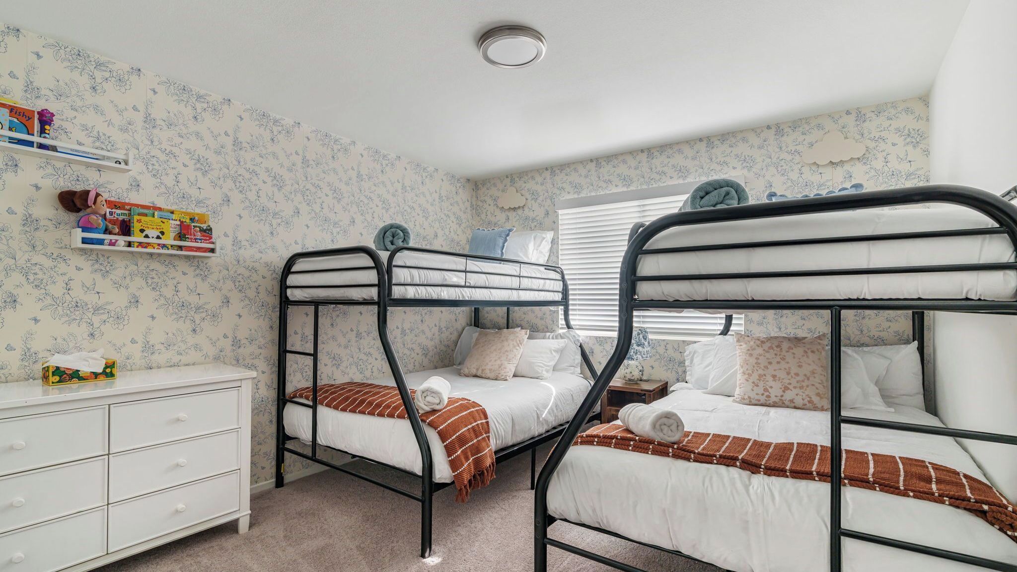 Een slaapkamer met stapelbedden, bloemenbehang, een witte dressoir en een plank vol kinderboeken.