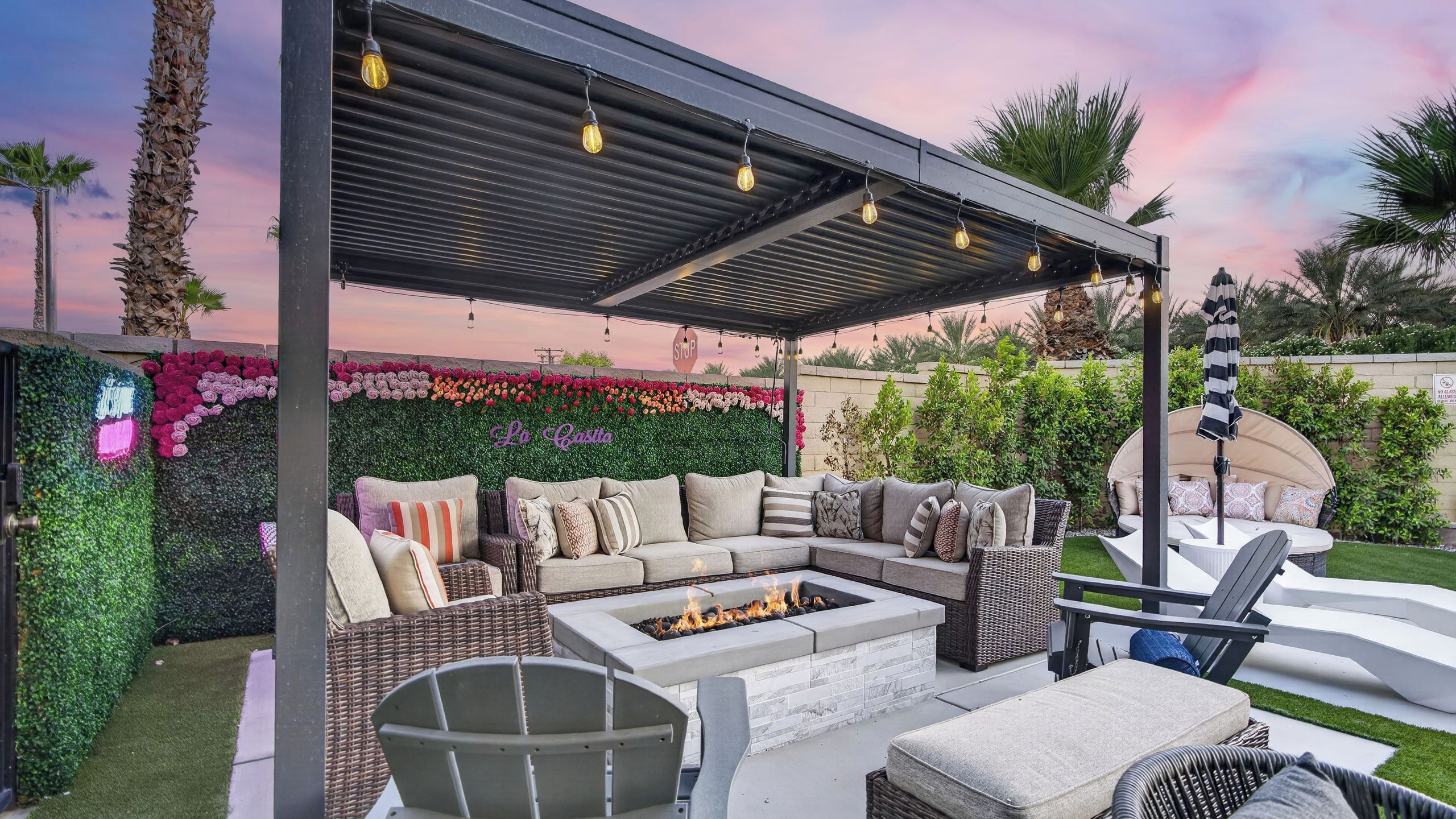A covered outdoor seating area with a fire pit, cushioned seating, and a pergola with string lights.