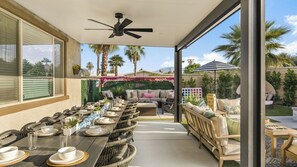 Un espace extérieur abrité, aménagé pour les repas, avec une longue table dressée, un ventilateur de plafond et une vue sur des palmiers et un jardin.