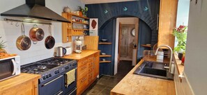 Eine Küche mit Herd, Backofen und hängenden Töpfen; ein Holzkabinett mit Geschirr; eine blaue Wand mit dekorativem Bogen; und ein Spülbecken mit Geschirrablage.
