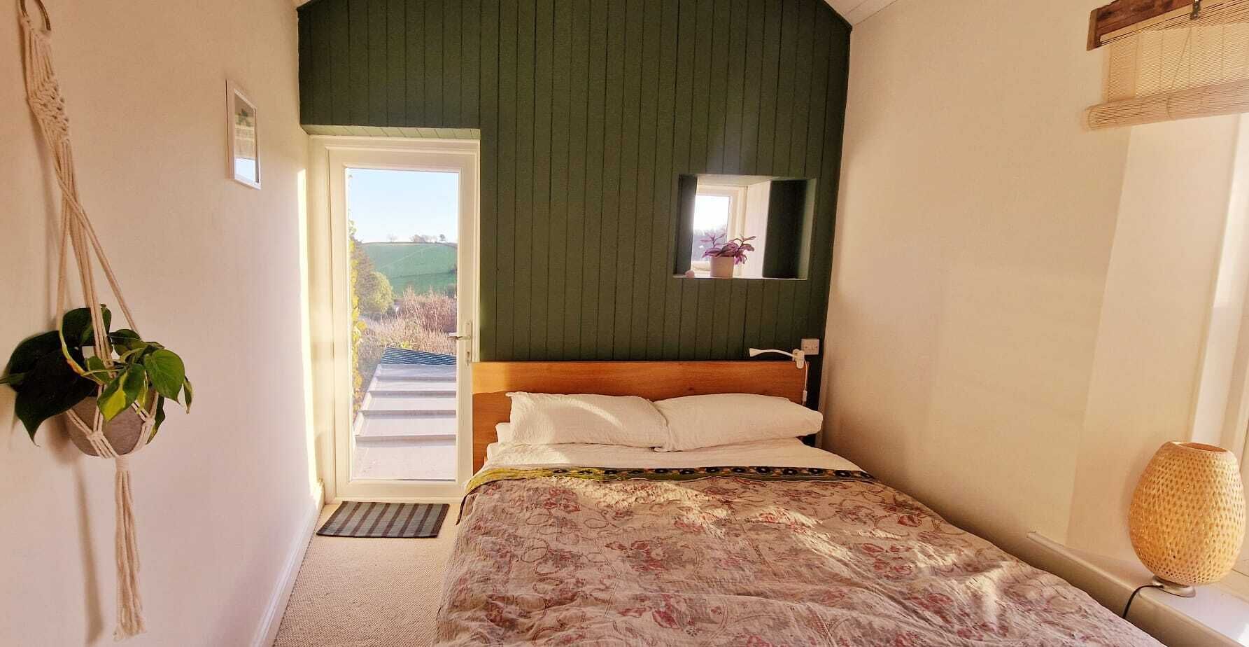 A bedroom with a bed, a small window, a macrame plant, and a view of the outdoors.