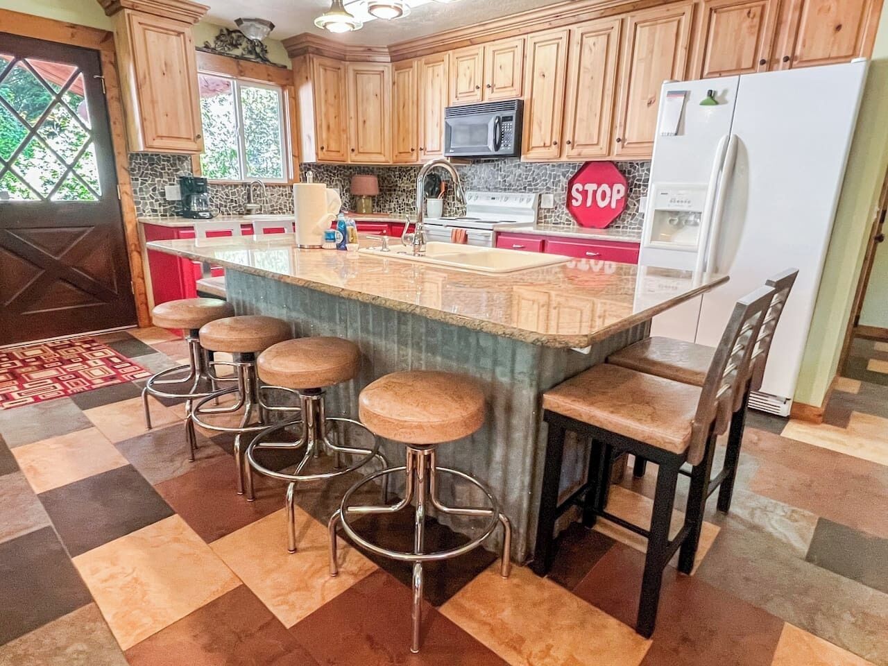 A kitchen with wooden cabinets, a granite countertop, a stainless steel sink, a microwave, a refrigerator, and a bar stool area.