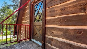 Una baita in legno con ringhiera in metallo rosso, una porta in legno decorata con un motivo a diamante e una vista aperta sul verde circostante.
