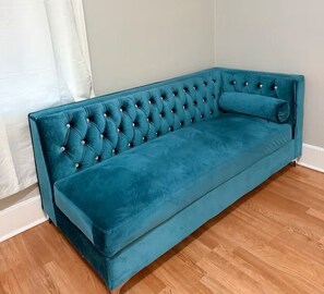 Ein smaragdgrünes Samtsofa mit gepolsterten Rückenlehne und Armlehnen, vor einer weißen Wand und Holzdielen.