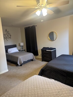 A bedroom with two beds, a ceiling fan, a nightstand with a lamp, a dresser, a mirror, and a decorative wall piece.