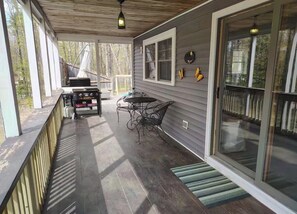 Eine überdachte Veranda mit Grill, Tisch und Stühlen sowie eine Glastür mit Blick auf Bäume.