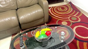 Een woonkamer met een leren bank, een glazen salontafel met een fruitschaal, een afstandsbediening en een kamerbreed tapijt.