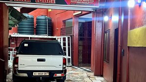 Een voertuig geparkeerd in een overdekte ruimte met een betegelde vloer, een bord erboven met de tekst 'O-ANSAH PLAZA LODGE', en een trap die naar een hoger niveau leidt.