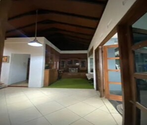 A hallway with a tiled floor, a ceiling with exposed beams, a hanging light fixture, and a door leading to a room with a green carpet. - Cozzy Pondok Sukamulus (Bandung Barat)