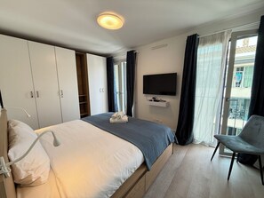 Ein Hotelzimmer mit Bett, einem an der Wand montierten Fernseher, einem Sessel und einem Balkon mit Vorhängen.