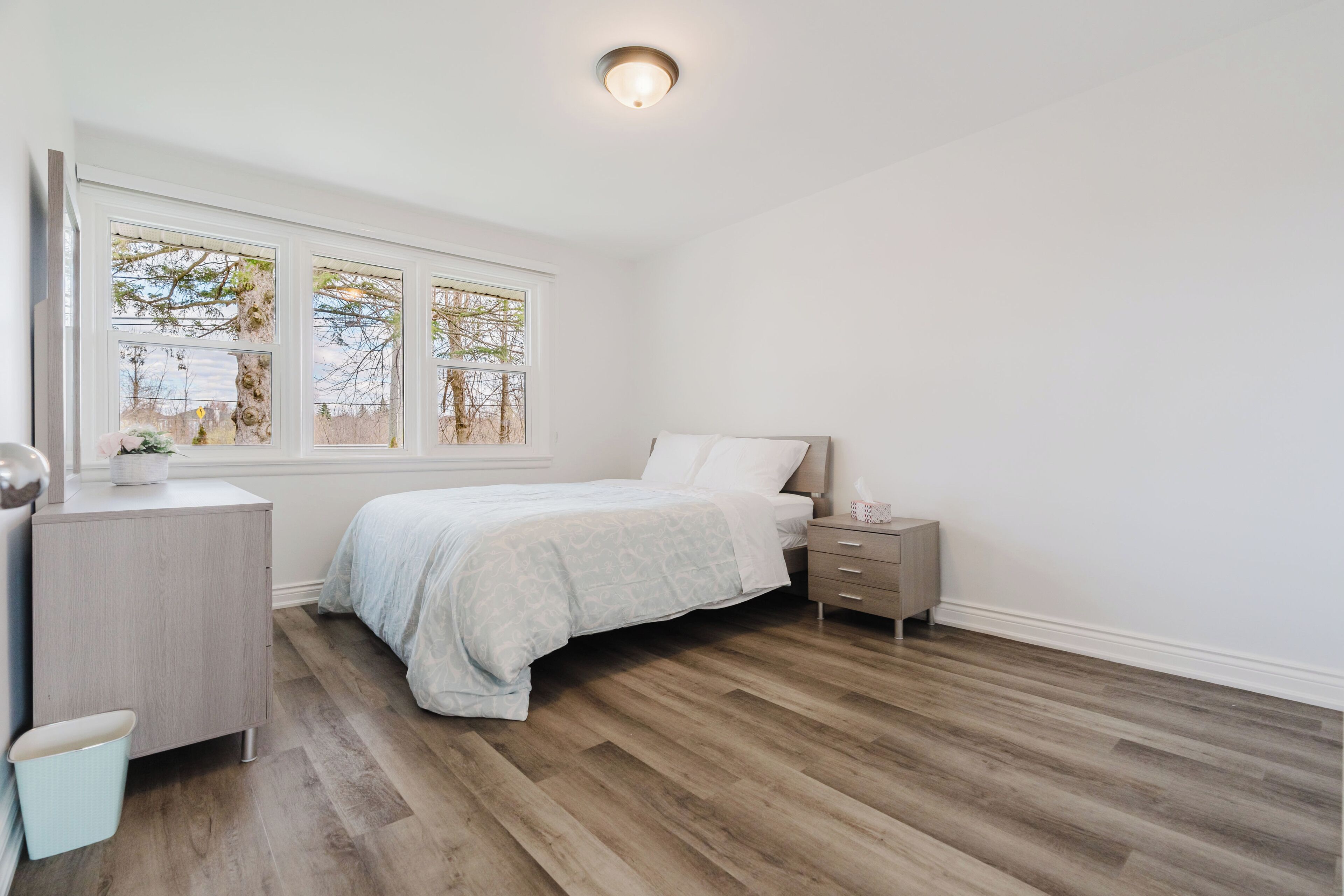 A bedroom with a bed, bedside table, nightstand, dresser, and a window with a view of trees.