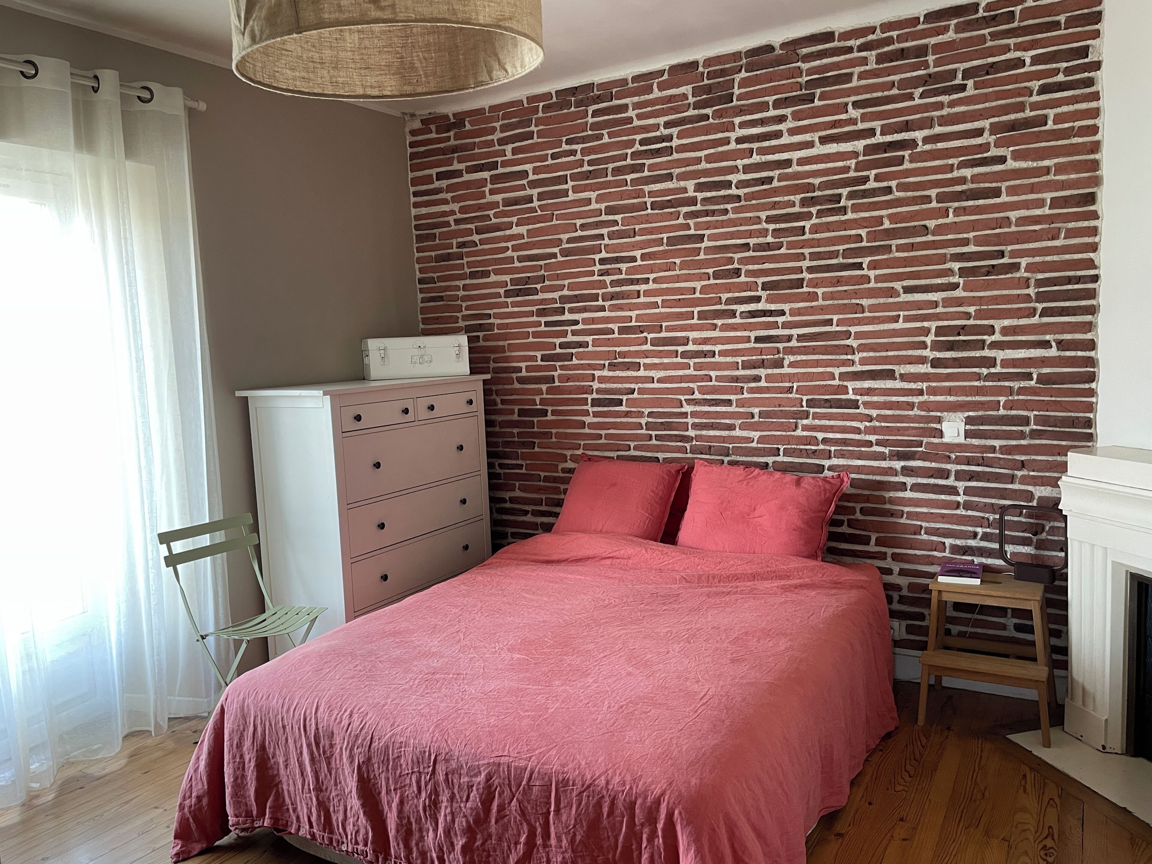 Une chambre avec un mur en briques, un lit avec une literie rose, une chaise en bois, une commode et un luminaire suspendu.