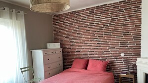 Et soveværelse med en murstensvæg, en seng med lyserødt sengetøj, en træstol, en kommode og en hængende lampe.
