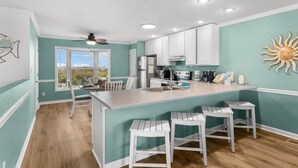 A kitchen with white cabinets, a central island with stools, a ceiling fan, and a view through a window. - The Mystic Turtle at Cape San Blas (Port St. Joe)