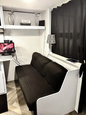 Et lille værelse med en sort sofa, et skrivebord med en computerskærm, en lampe og et vindue med sort gardin.