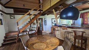 Un interior rústico con una escalera de madera, una mesa redonda de madera para comer con individuales tejidos, y una zona de cocina con taburetes de bar.